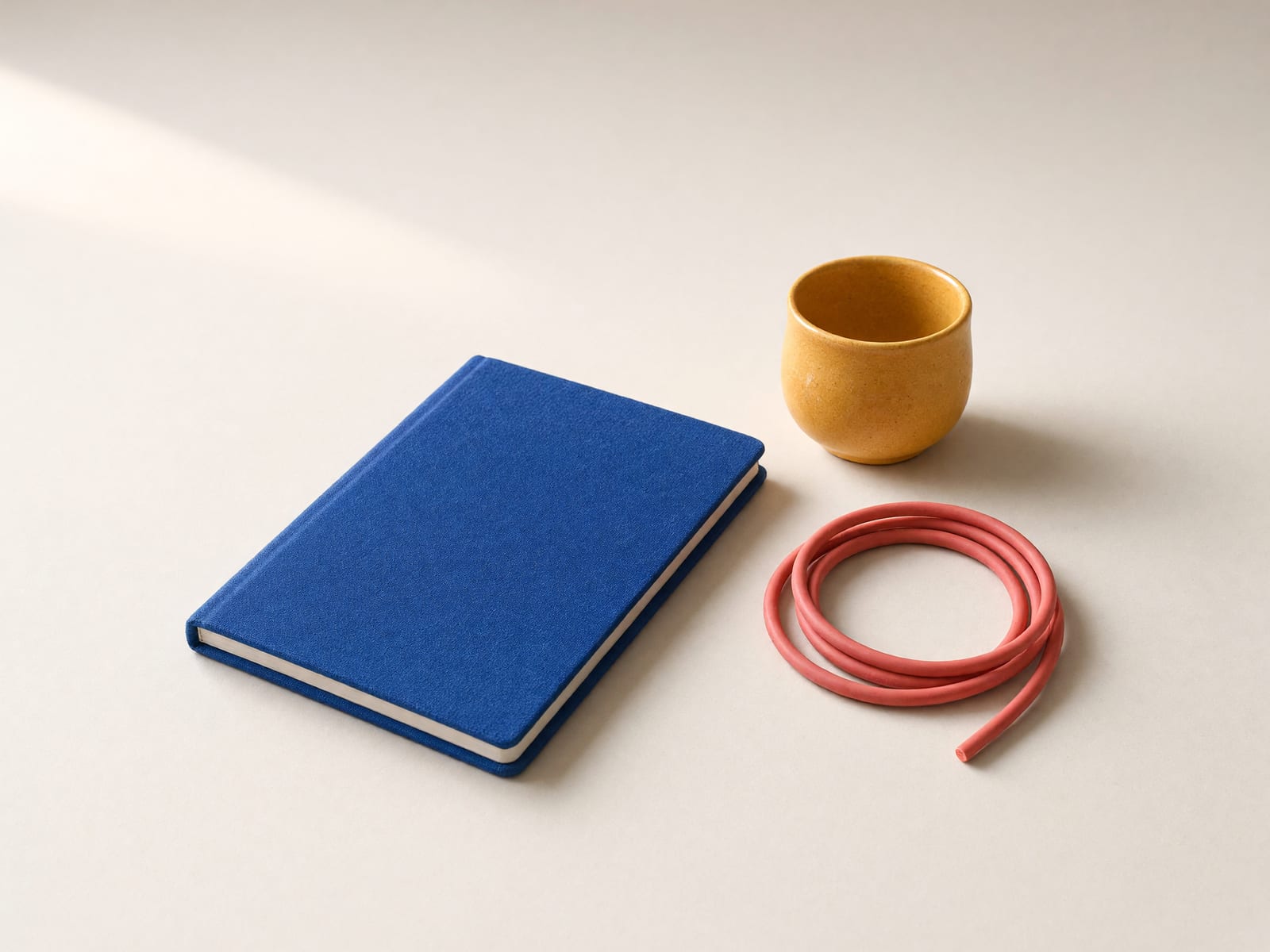 Geschlossenes blaues Notizbuch, ein locker aufgewickeltes korallrotes Kabel und eine kleine gelbe Tasse auf hellem Untergrund.