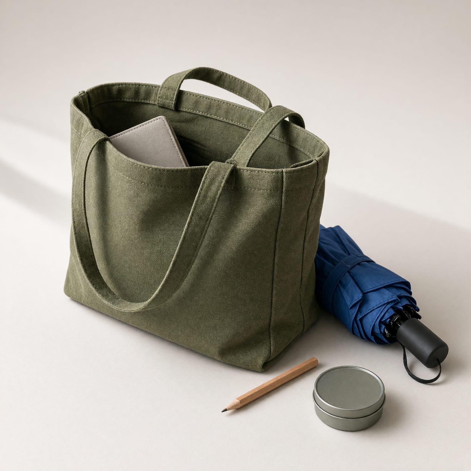 Offene grüne Leinwandtasche mit einem gefalteten Schirm, einem kurzen Holzstift und einer kleinen runden Dose daneben.
