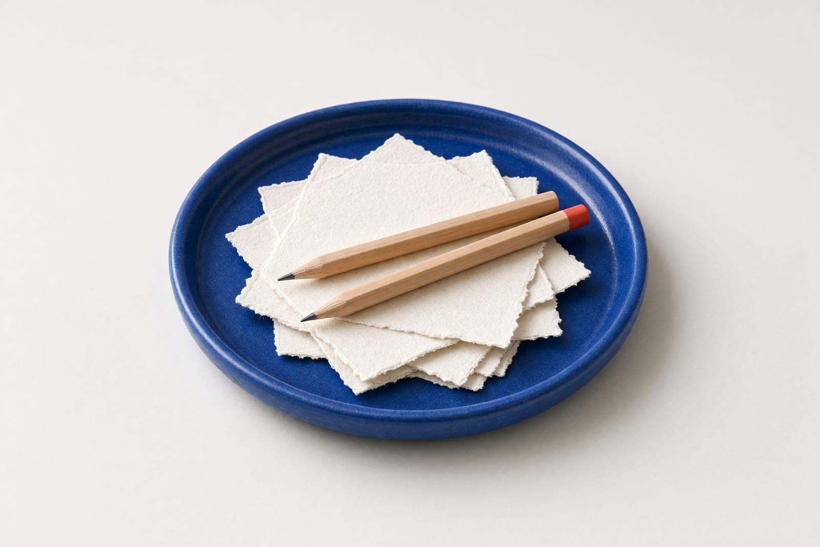 Eine flache blaue Keramikschale mit losen Papierstreifen und zwei Holzstiften auf hellem Untergrund.