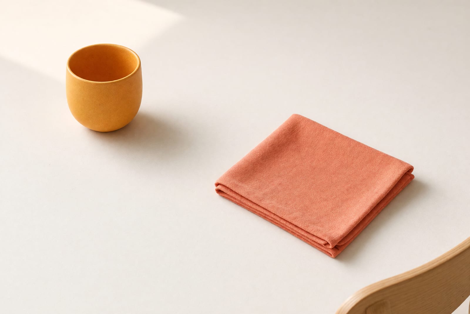 Fast leere Tischfläche mit einer gelben Tasse und einem gefalteten korallroten Stoff, rechts der Rand einer Stuhllehne.